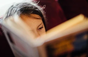 A young child engrossed in reading a book, with their face partially hidden behind the pages.