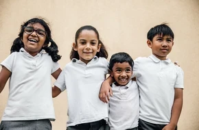 Four smiling primary school children wearing white polo shirts stand together against a beige wall, with their arms around each other.